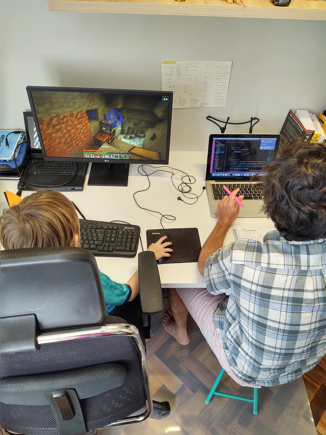 A picture from behind, showing my son playing minecraft on a big display with a nice chair, and I working on small stool on a small screen laptop.